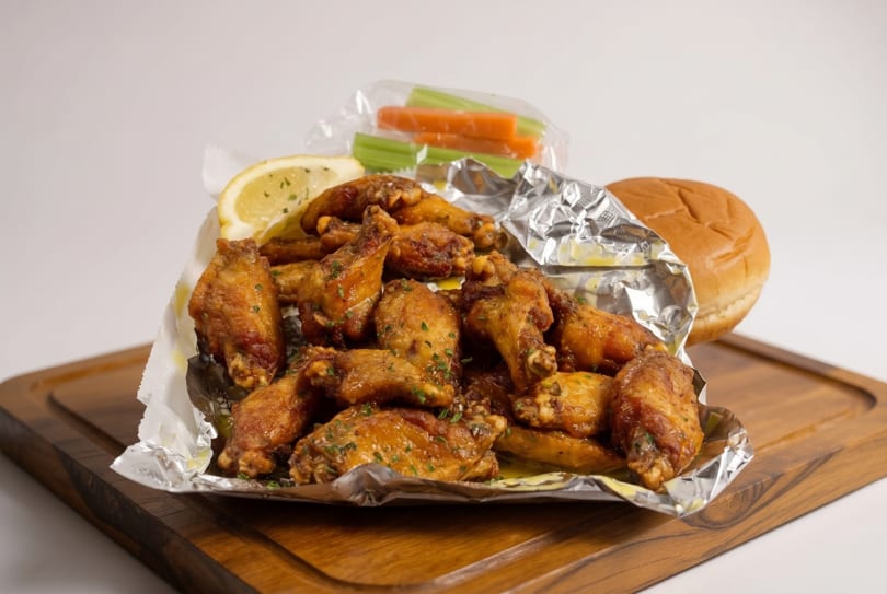 Crispy chicken wings in foil with a lemon slice, celery, carrots, and a bun on a wooden board.