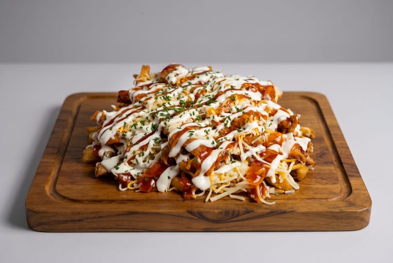 Loaded fries with chicken, cheese, white sauce, BBQ sauce, and parsley on a wooden board.