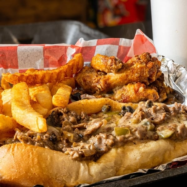 Cheesesteak sub with fries and chicken wings in a black tray with red and white checkered paper.