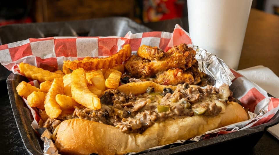 Cheesesteak sub with fries and chicken wings in a black tray with red and white checkered paper.