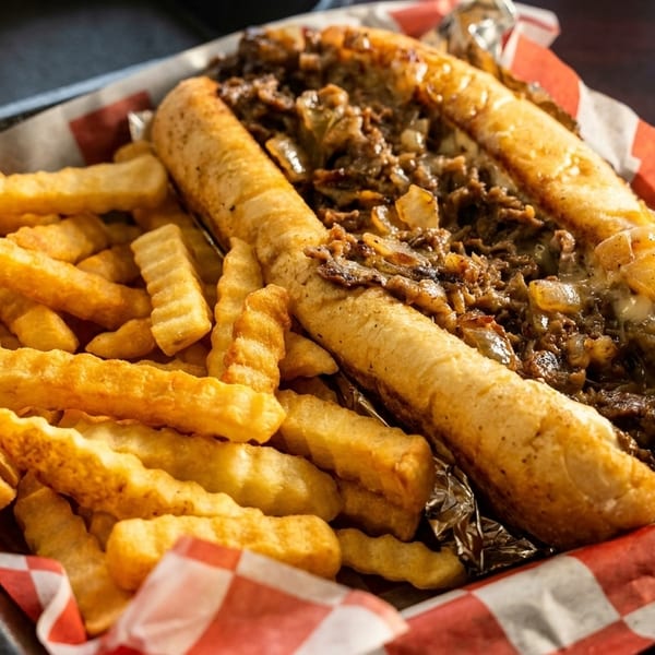 Cheesesteak sandwich with melted cheese and onions, served with crinkle-cut fries in a basket.