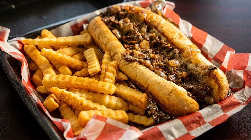 Cheesesteak sandwich with melted cheese and onions, served with crinkle-cut fries in a basket.