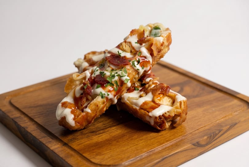 Two crispy waffle sticks topped with white sauce, BBQ sauce, bacon, and parsley on a wooden board.