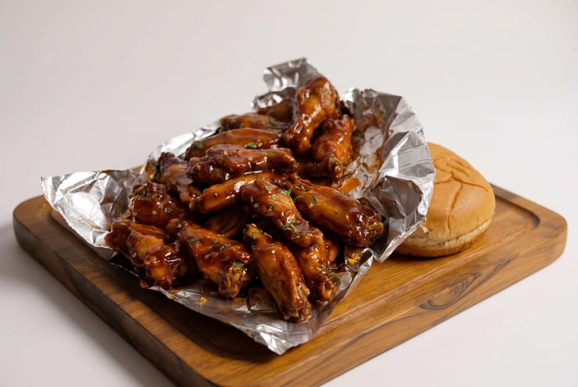 A pile of glistening, saucy chicken wings in foil on a wooden board with a burger bun.