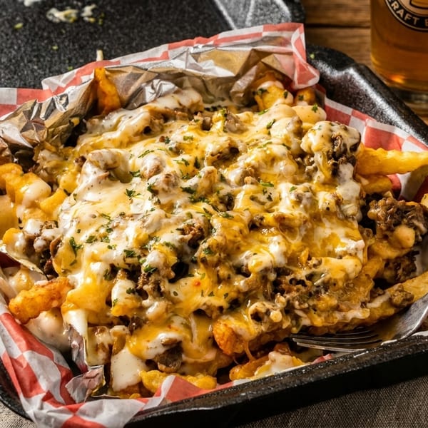 Loaded fries with melted cheese, ground meat, and herbs in a takeout container with a beer and hot sauce.