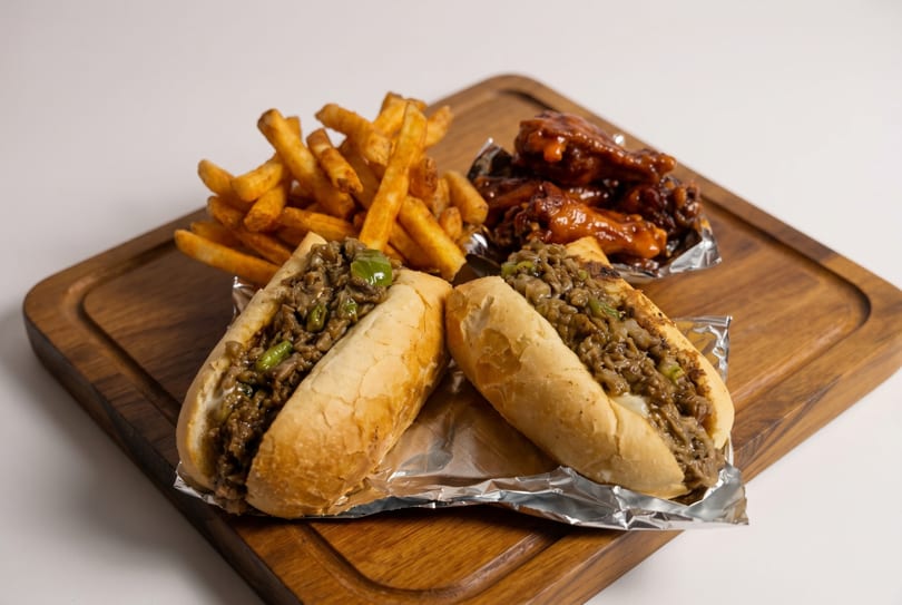 Two cheesesteak sandwiches, a pile of golden fries, and BBQ chicken wings on a wooden board.